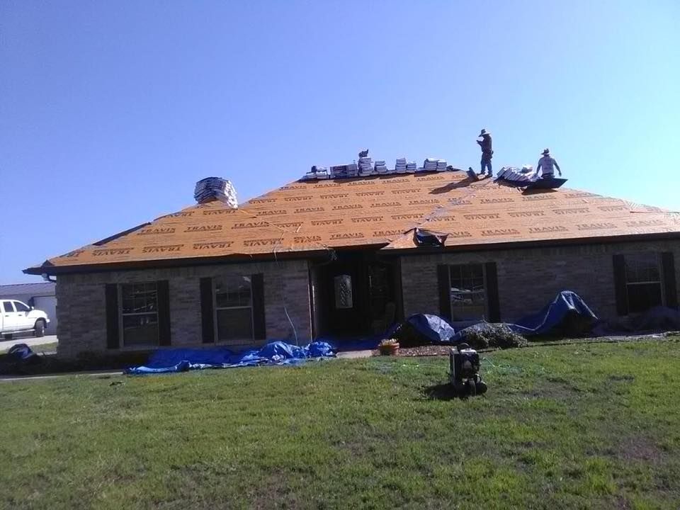 Workers replacing roof shingles on a brick house under a blue sky; tarps on ground.