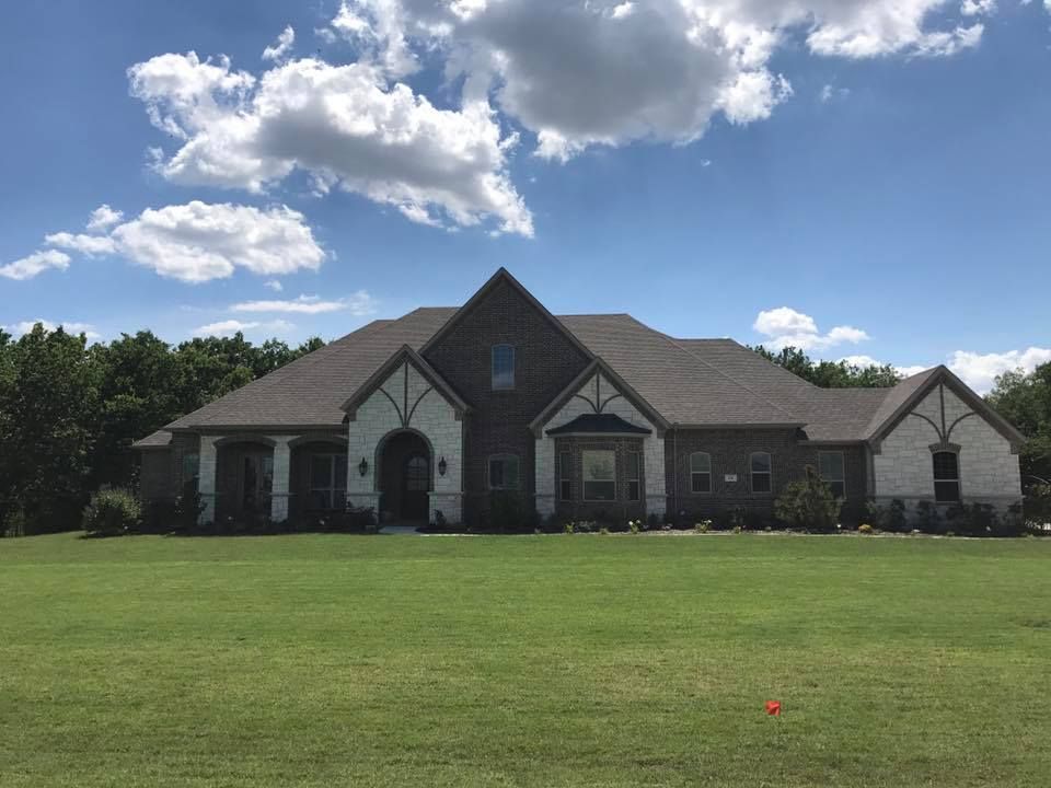 A large house with brown roof and brick and stone exterior, set against a blue sky with clouds.