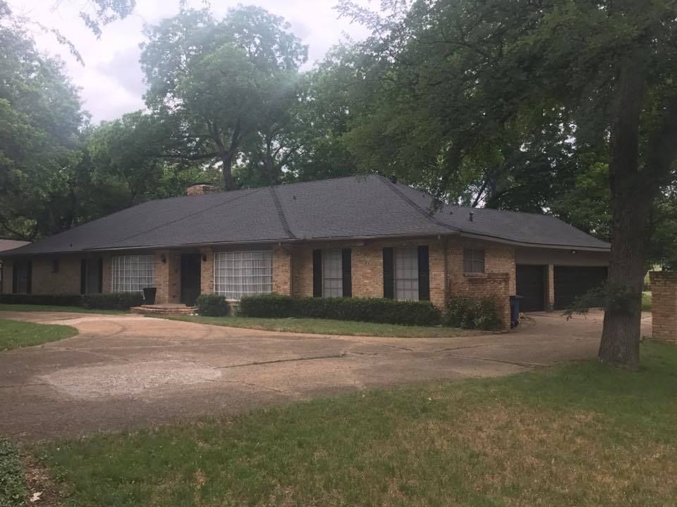 Brick ranch-style house with dark roof and attached carport, surrounded by trees and grass, on a cloudy day.