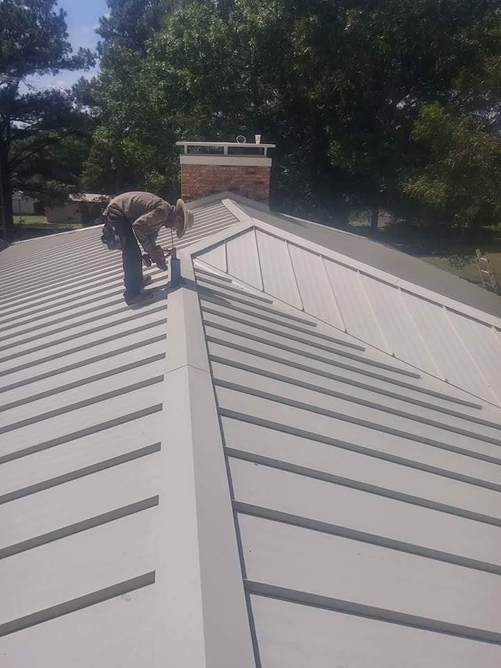 Person working on a gray metal roof, near a brick chimney. Trees in the background.