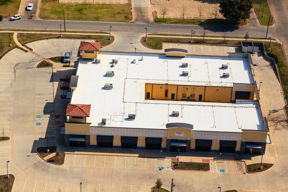 Aerial view of a commercial building with a flat white roof, parking lot, and surrounding streets.
