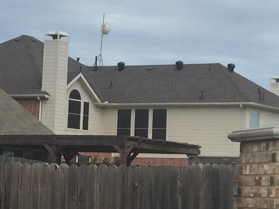 Two-story beige house with dark roof and chimney, seen over a wooden fence and pergola, under a cloudy sky.