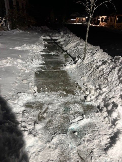 A snowy sidewalk with a tree in the background at night.