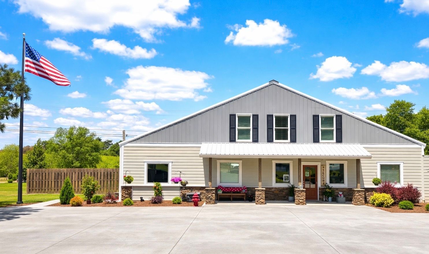 A light-colored building with a gray roof and a front porch under a blue sky, with an American flag flying on the left.