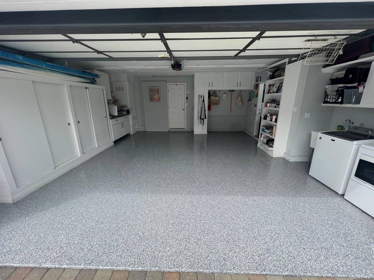 A clean, empty garage with a grey speckled epoxy floor, white storage cabinets, and shelving units along the walls.