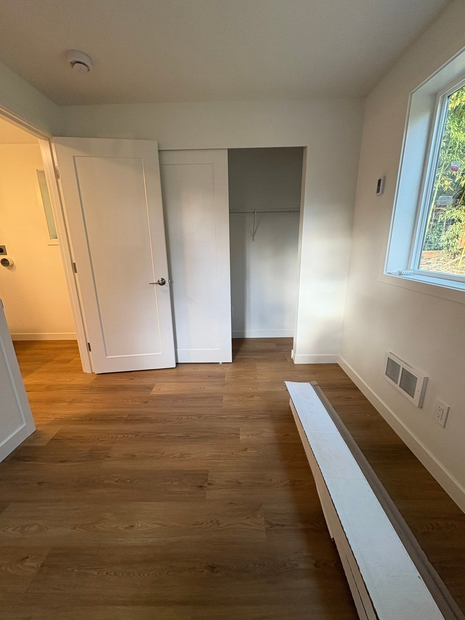 An empty room with light wood floors, white walls, a white interior door, a small walk-in closet, and a heating vent.