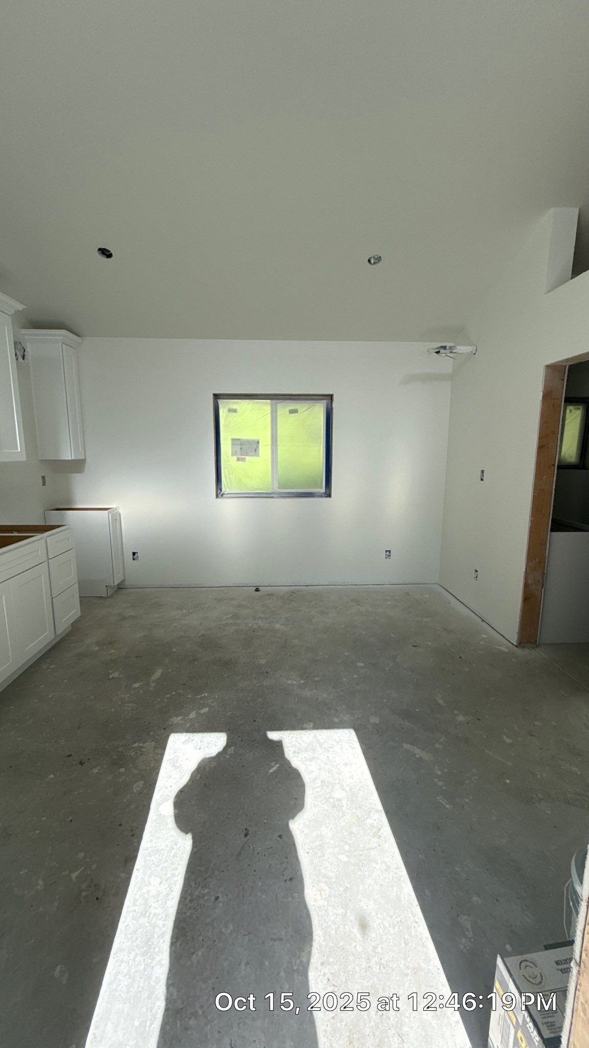 An unfinished room with white walls, gray concrete flooring, a central window, and white kitchen cabinets on the left.