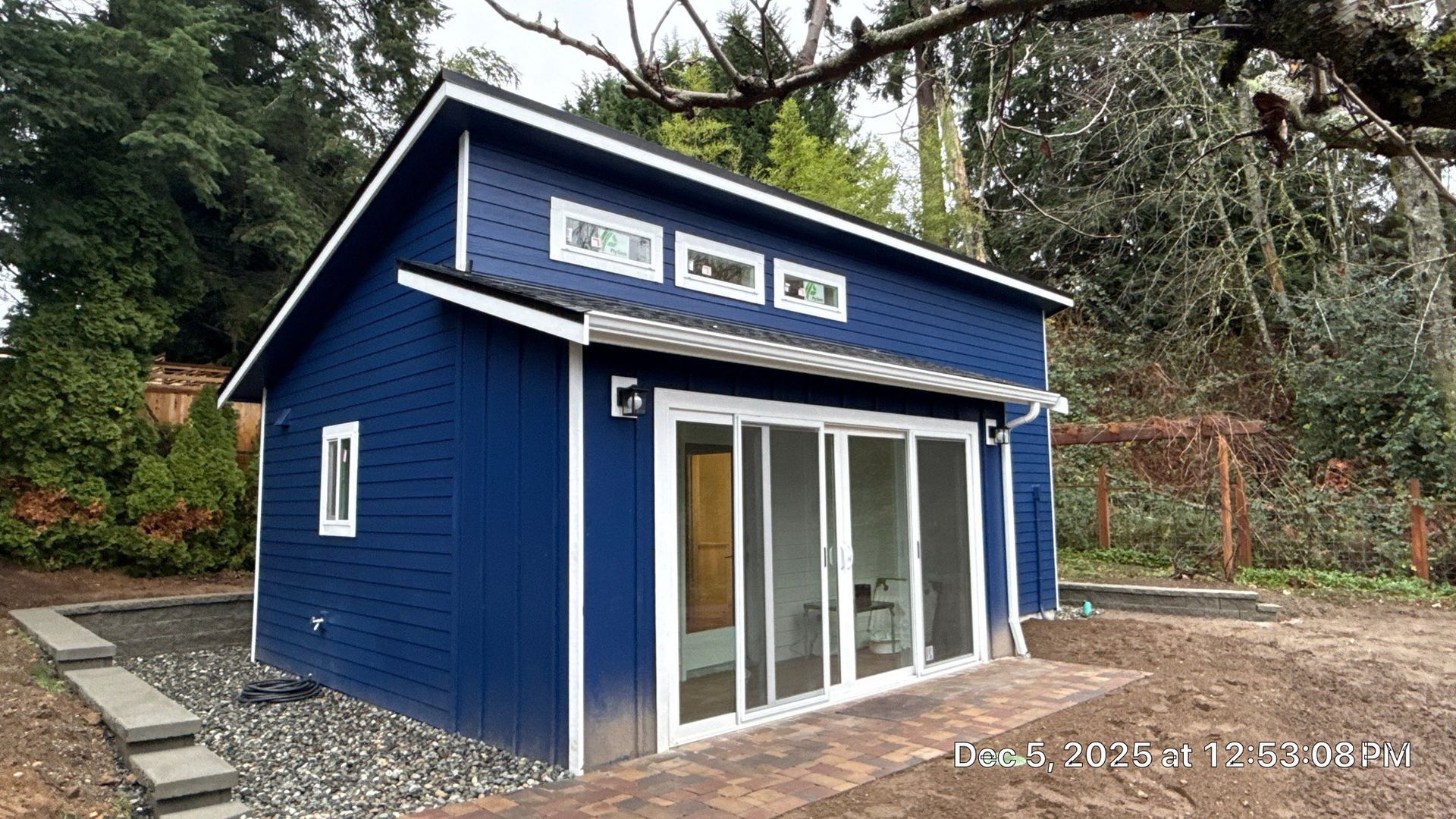 A small, deep blue modern shed with white trim, large sliding glass doors, and clerestory windows in a wooded backyard.