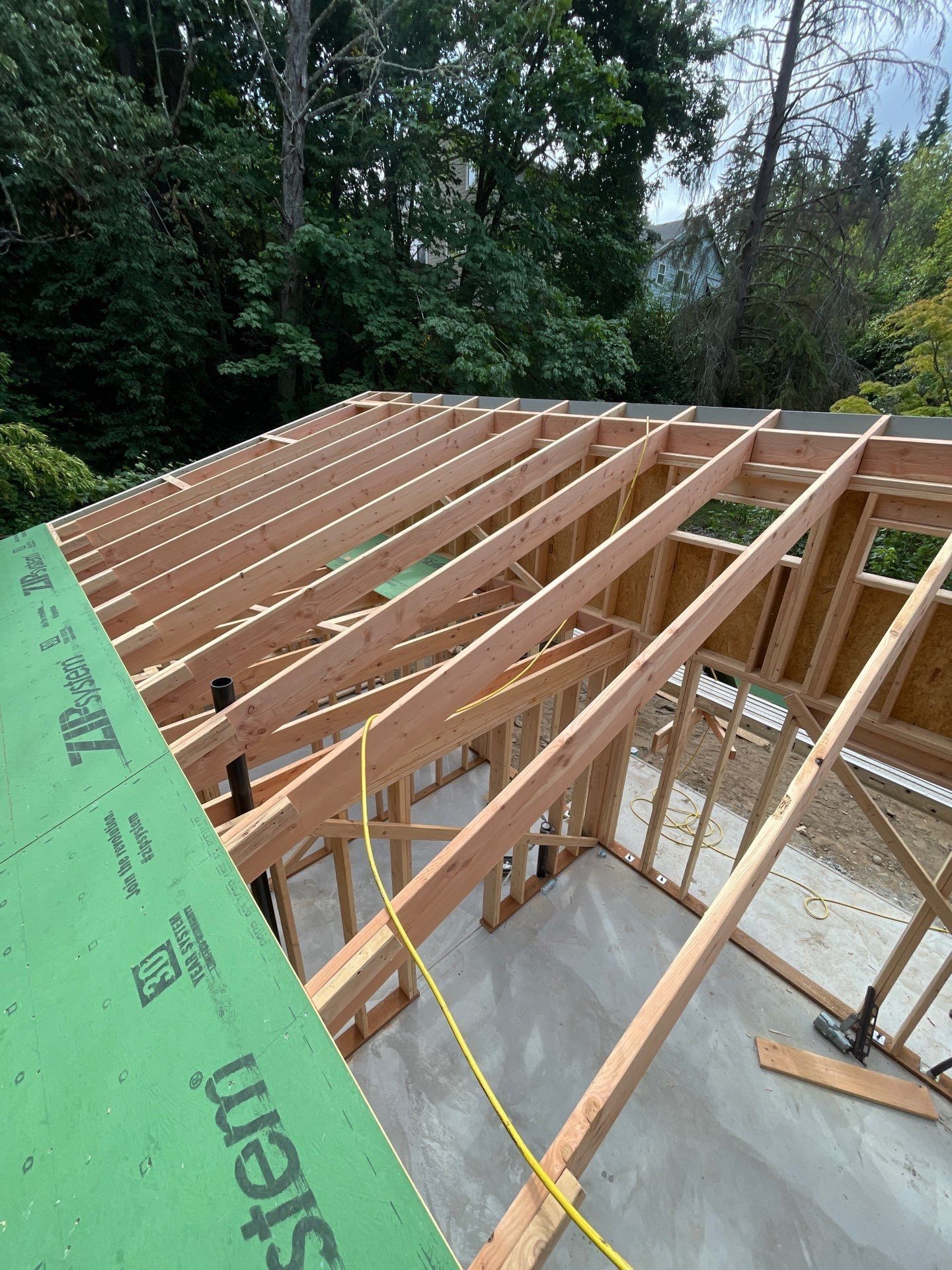 A construction site showing a concrete foundation, a wooden wall frame, a blue ladder, and stacks of lumber.