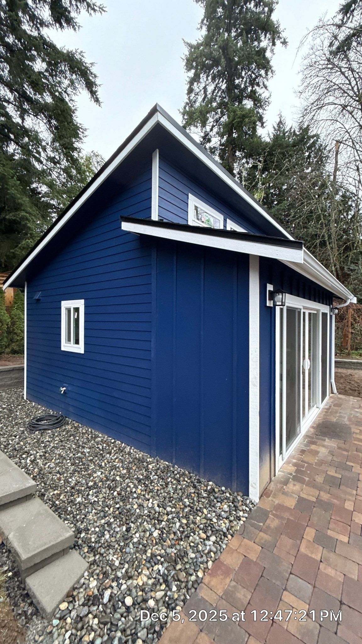 A blue shed with a white roof trim and window, set on a gravel base next to a brick patio with stairs.