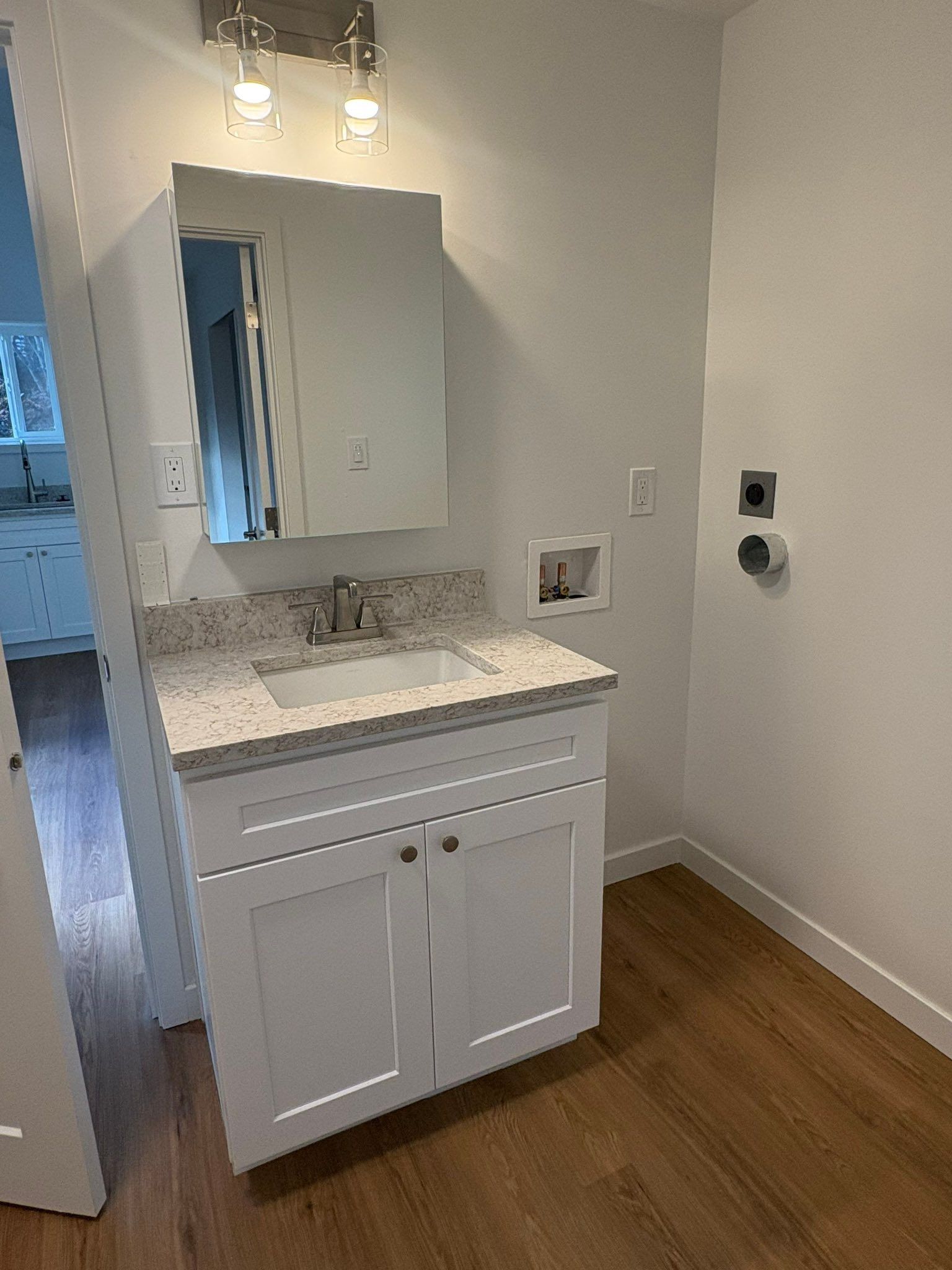 A bathroom vanity with a white cabinet, speckled countertop, sink, and mirror, next to laundry hookups on a wood floor.
