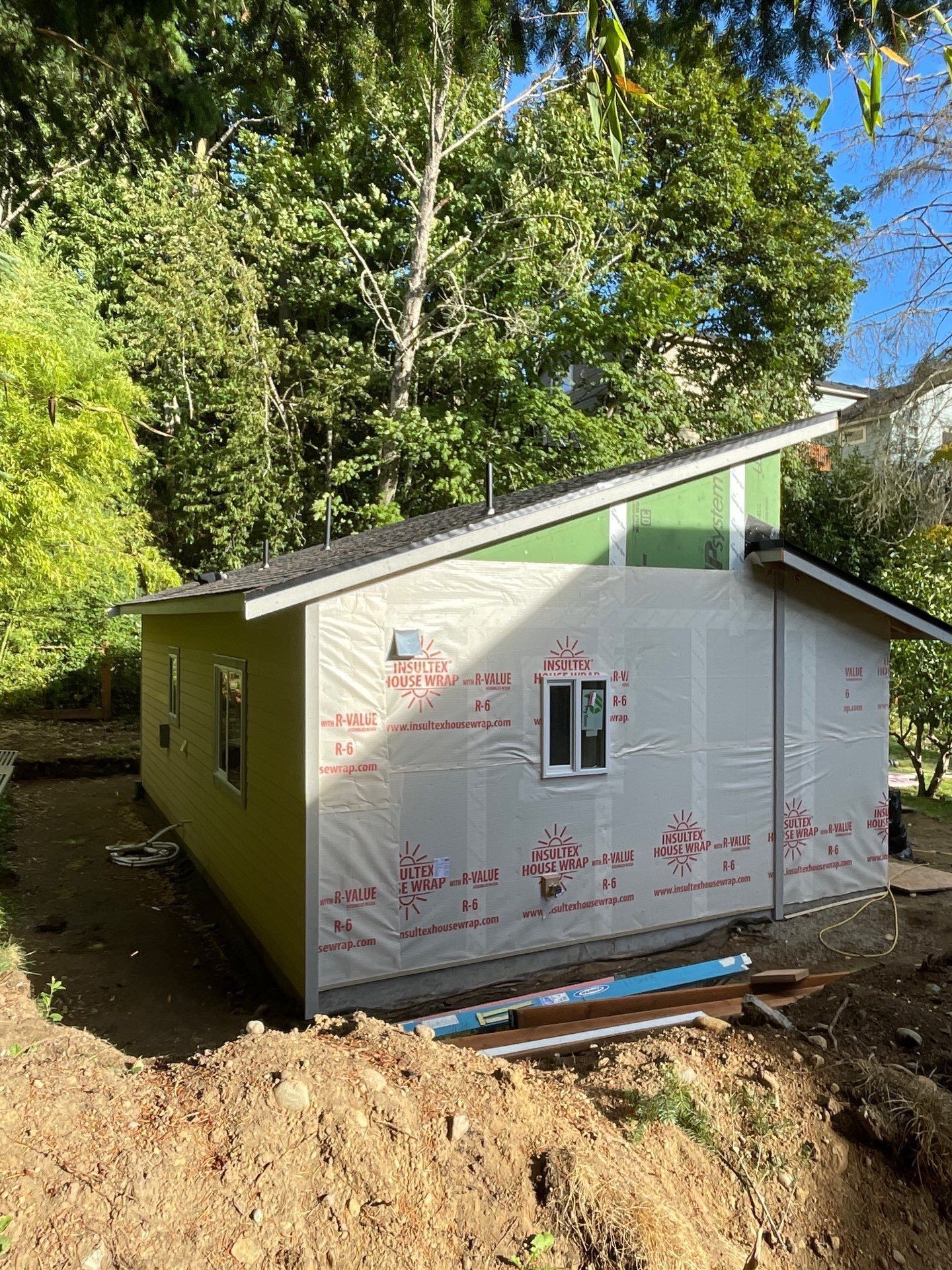 A partially constructed shed with white house wrap, a shed roof, a small window, and green trim, set in a wooded area.