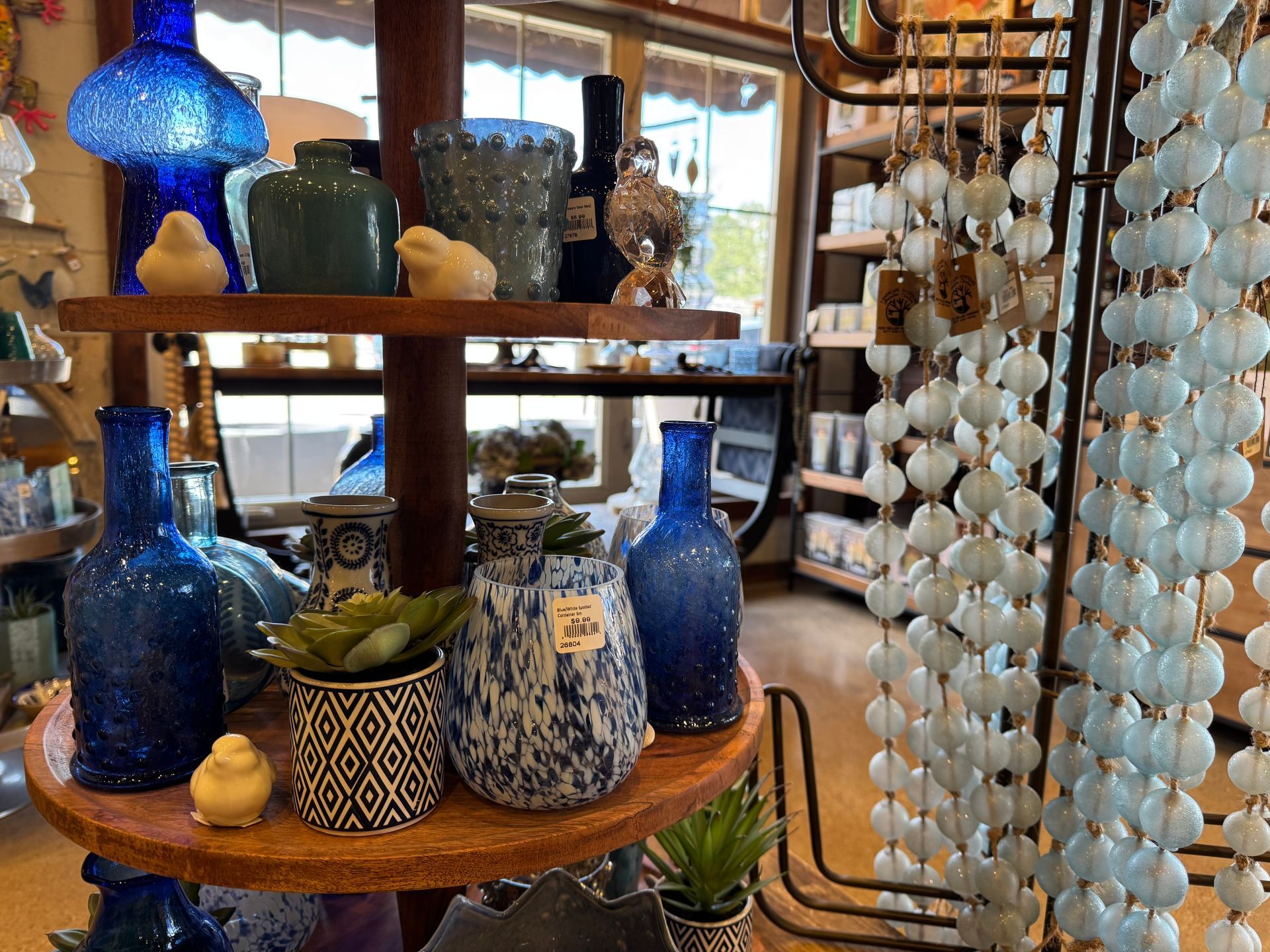 Blue glass vases (various shades and textures), Greenish-blue ceramic vase, Patterned ceramic pots (with and without faux succulents), White ceramic bird figurines, Round wooden tiered display stand, Hanging beaded garlands (light blue/white color)
Small decorative jars and bottles
Faux succulents in patterned planters.