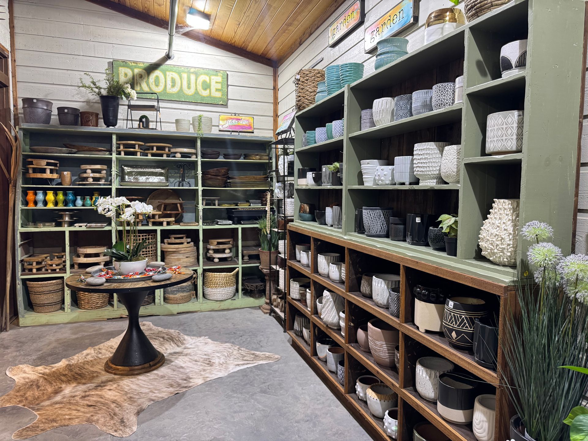 Assorted decorative pots and planters (ceramic, textured, patterned, and neutral tones), Wooden risers and trays (various sizes and shapes), Woven baskets (stored on lower shelves), Mini vases and colorful bud vases (including bright orange, blue, and green), Tabletop display with orchids, small plants, and mini gift items, Cowhide rug, Tall faux stems and greenery, Vintage green