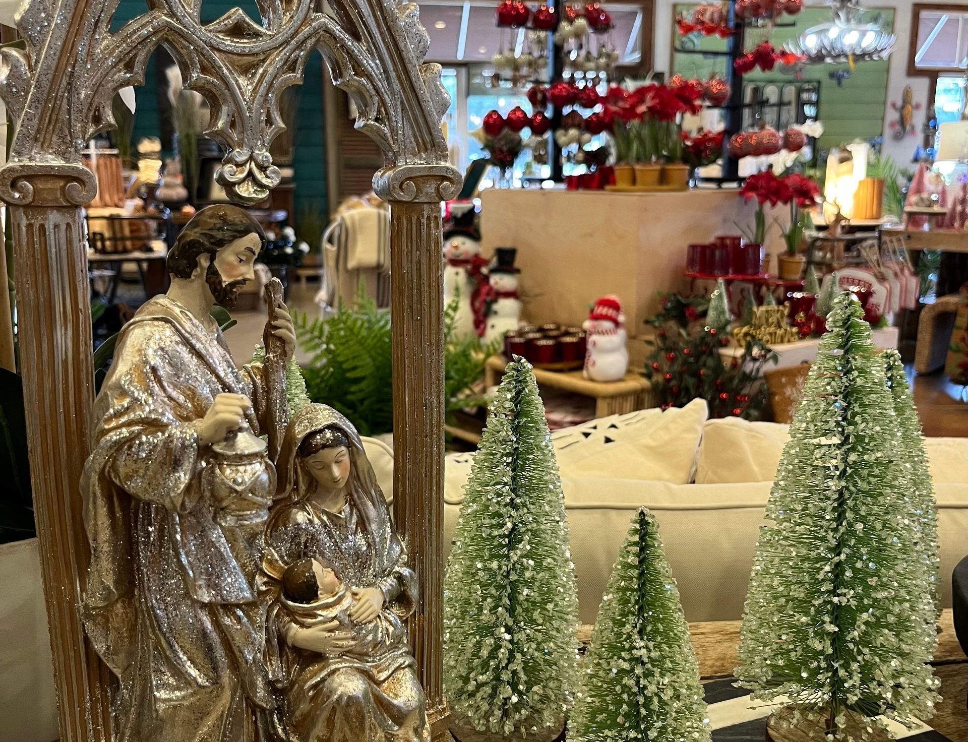 Nativity depicting the Birth of Jesus and sparkling green tabletop Christmas trees in the foreground, with red and white Christmas decor in the background.