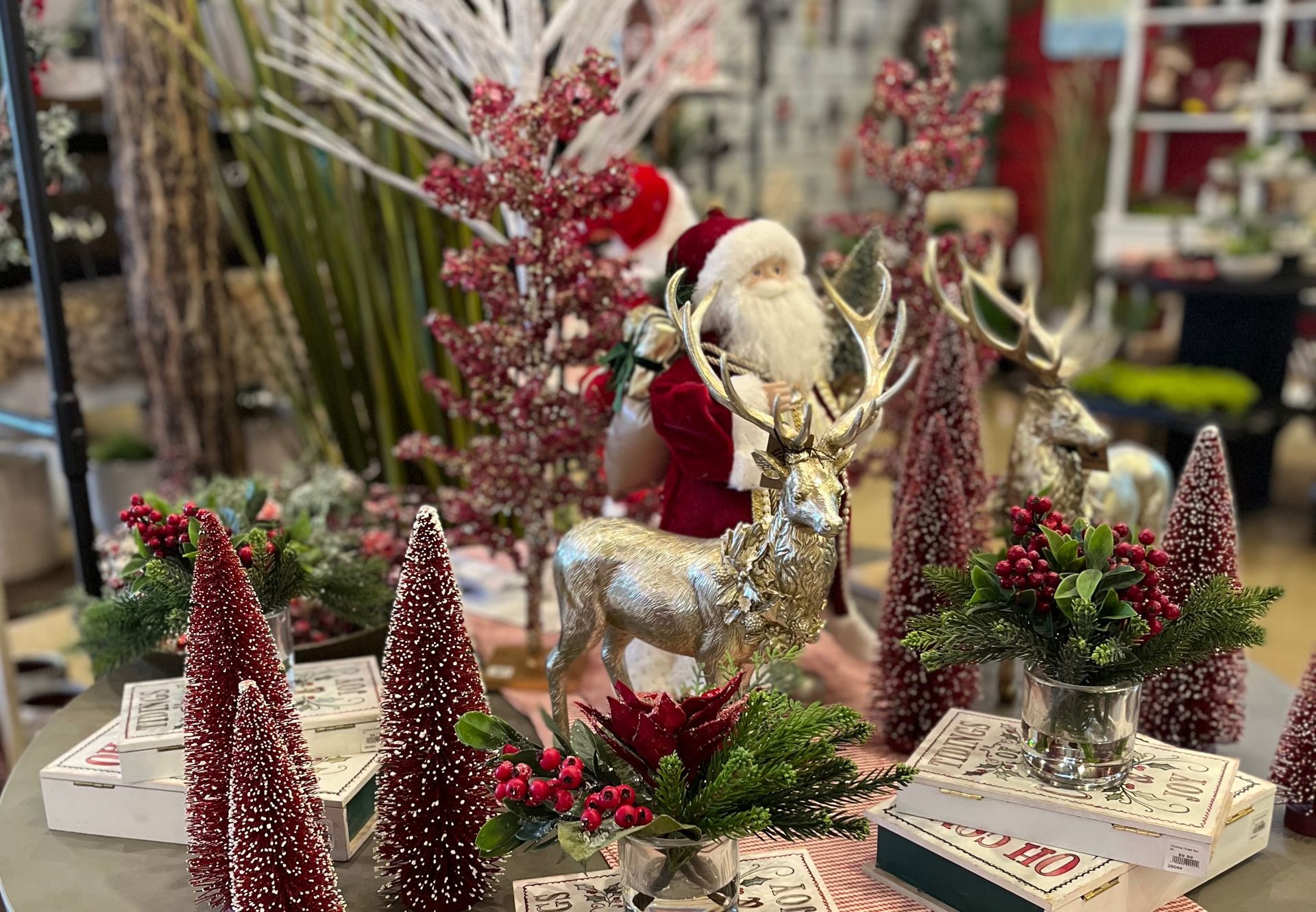 Table top Chrsitmas decor: red glitter trees, Santa Claus, books, metallic reindeer, Holly berries, and greenery.