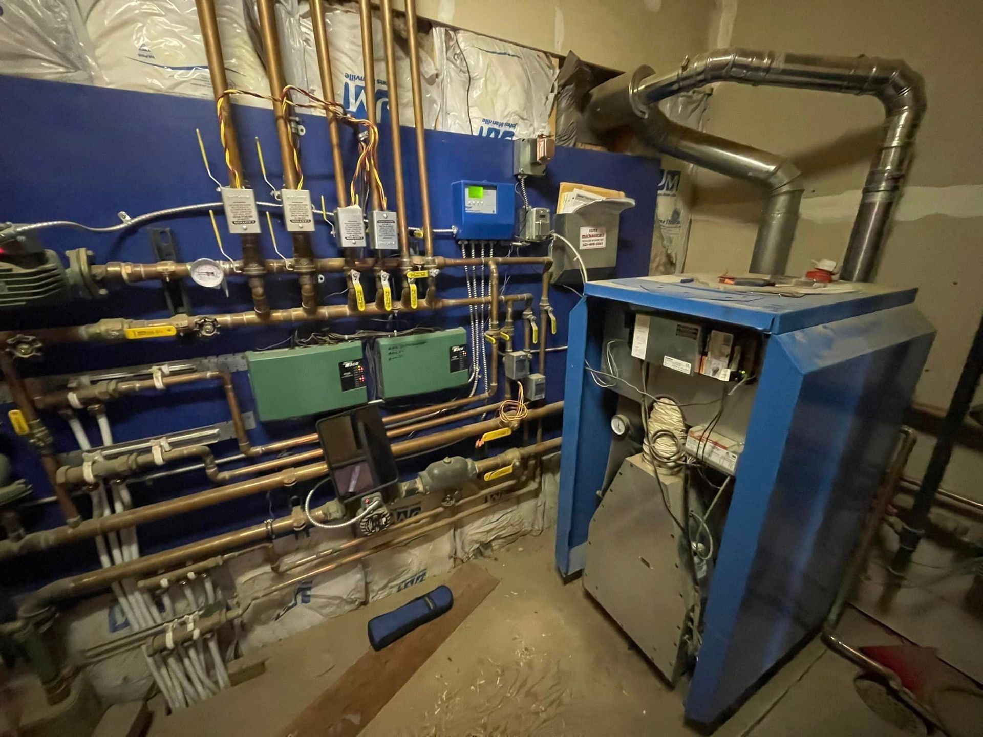 Blue boiler with open panel, pipes, gauges, and electrical components on a blue wall in a utility room.