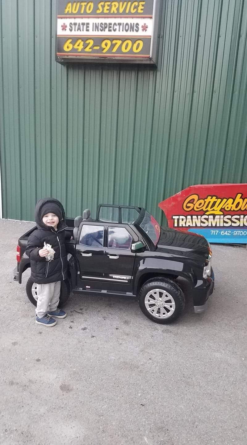 Young child stands by a black toy SUV in front of an auto repair shop with green siding.