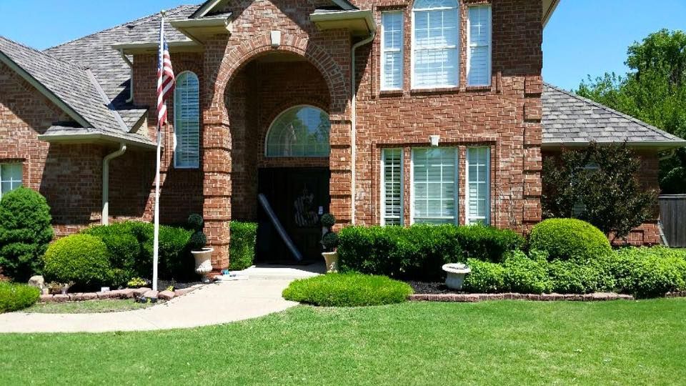 Brick house with arched entrance, American flag, and well-manicured lawn. Green shrubs and grass create a tidy curb appeal.