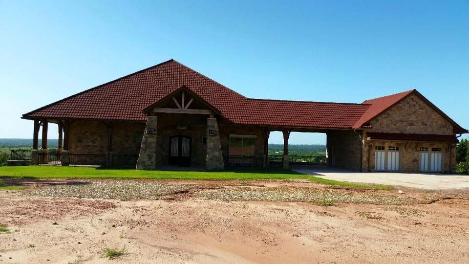 Stone house with a red tiled roof and a carport. The house is on a slightly elevated plot of land under a clear blue sky.