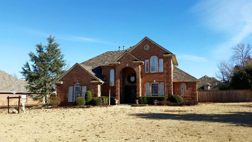 Brick house with brown roof, shutters, and a front yard, under a blue sky.