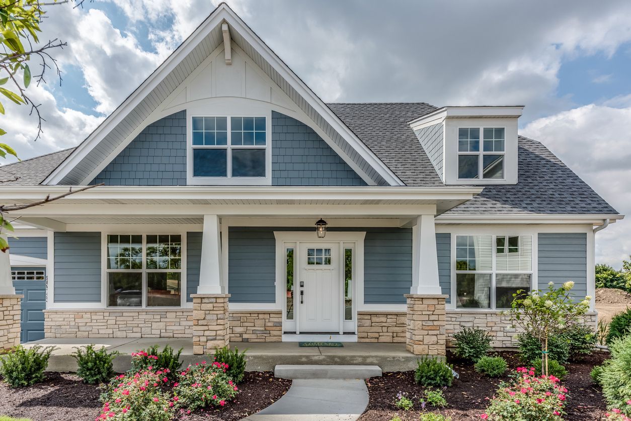 Blue and white craftsman-style house with stone accents, covered porch, and a landscaped front yard under a cloudy sky.