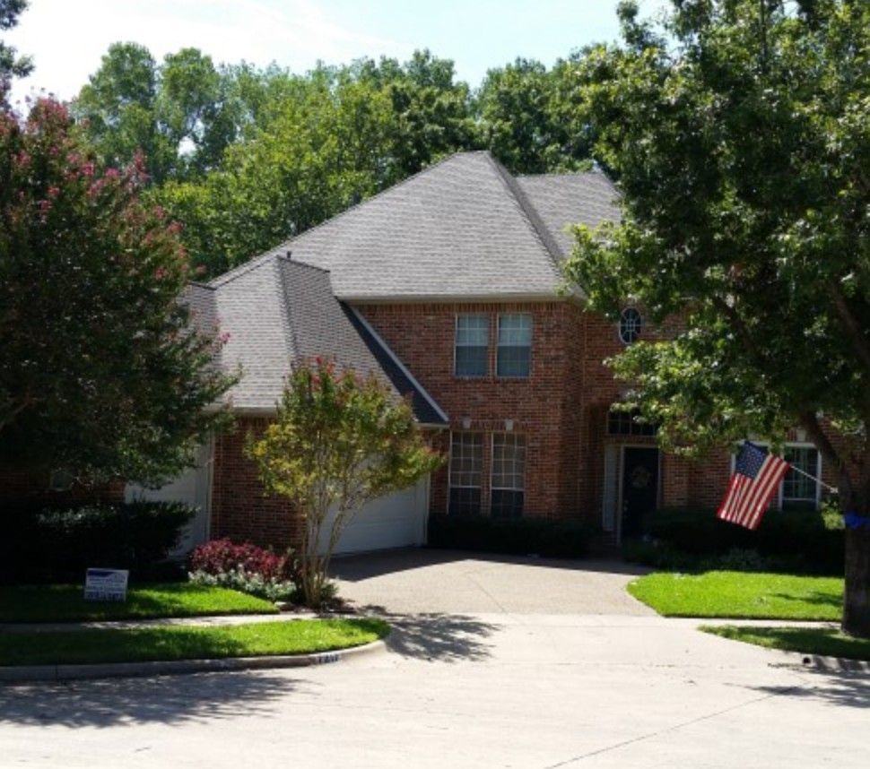 Two-story brick house with a driveway and an American flag. Trees surround the house on a sunny day.