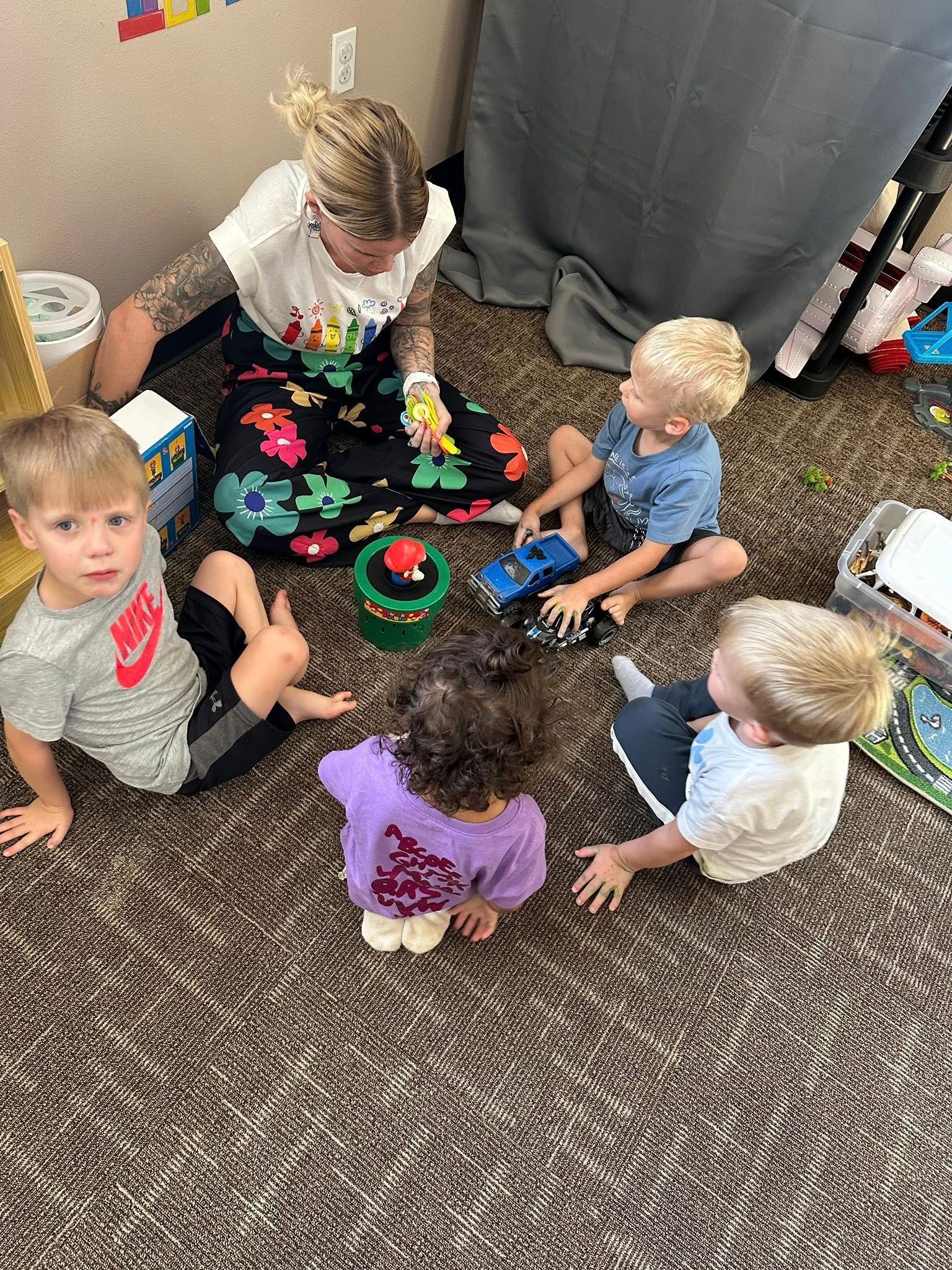 A woman and four toddlers sit on the floor. They play with toys indoors.