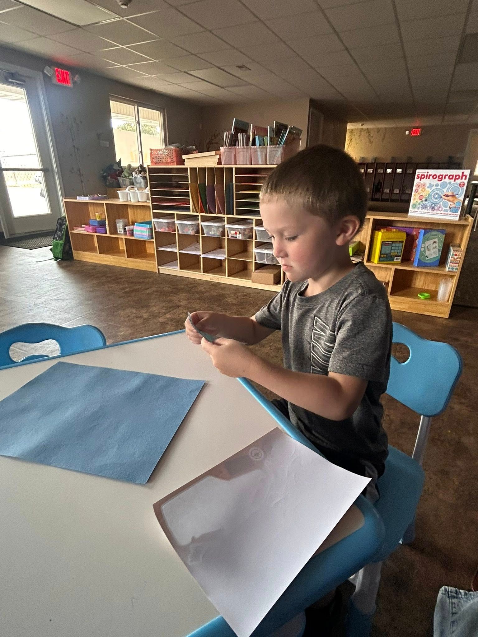 Young boy seated at table, focused on crafting with paper in classroom.