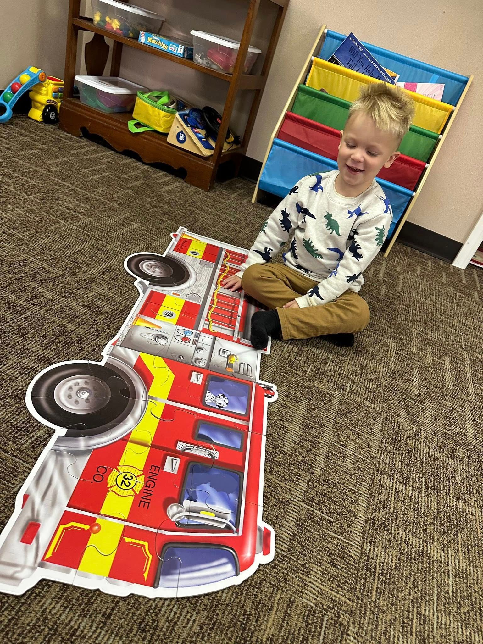 Young boy smiles while assembling a large fire truck puzzle on the floor.