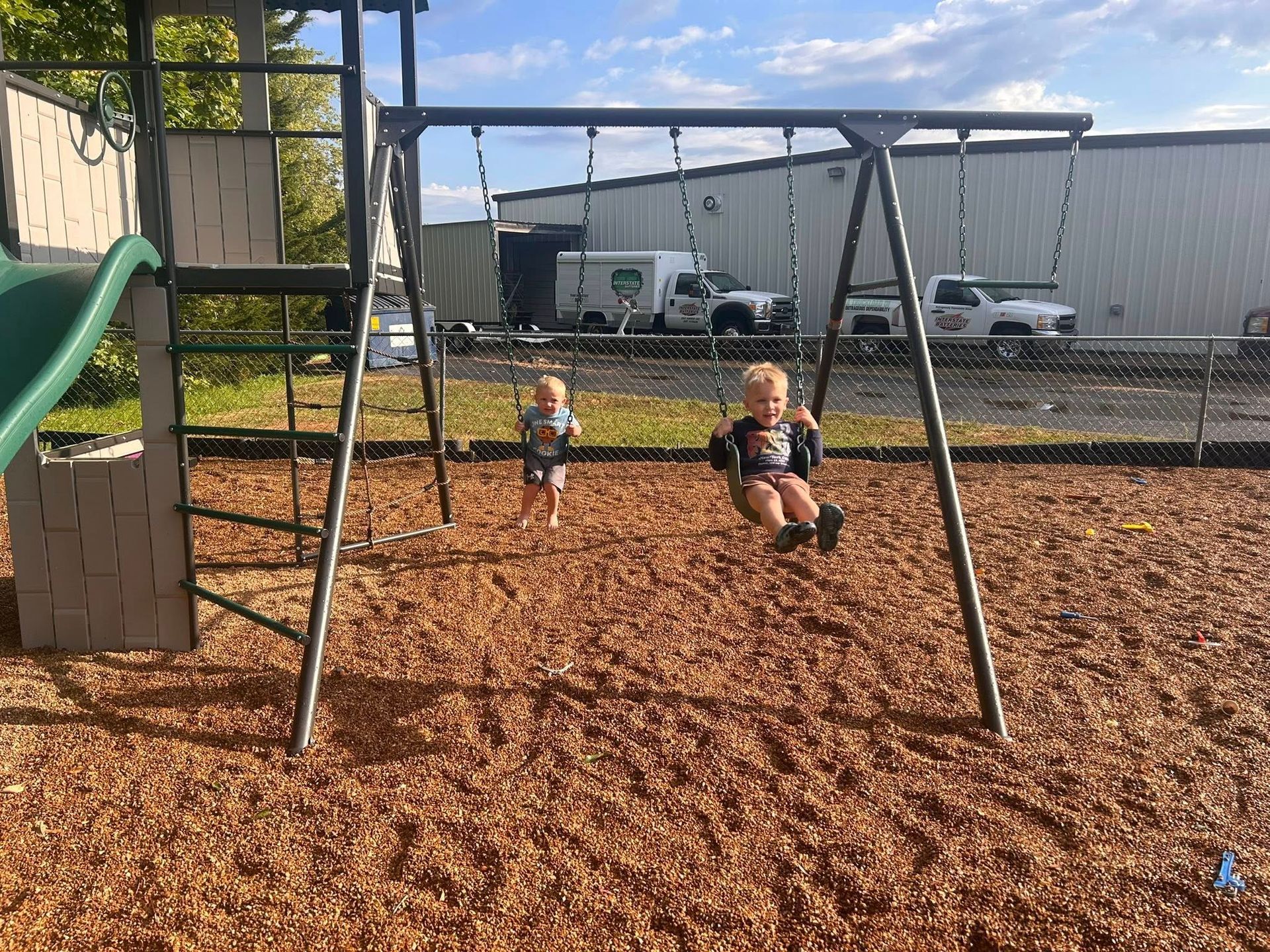 Two young children swinging at a playground. One is swinging higher than the other.