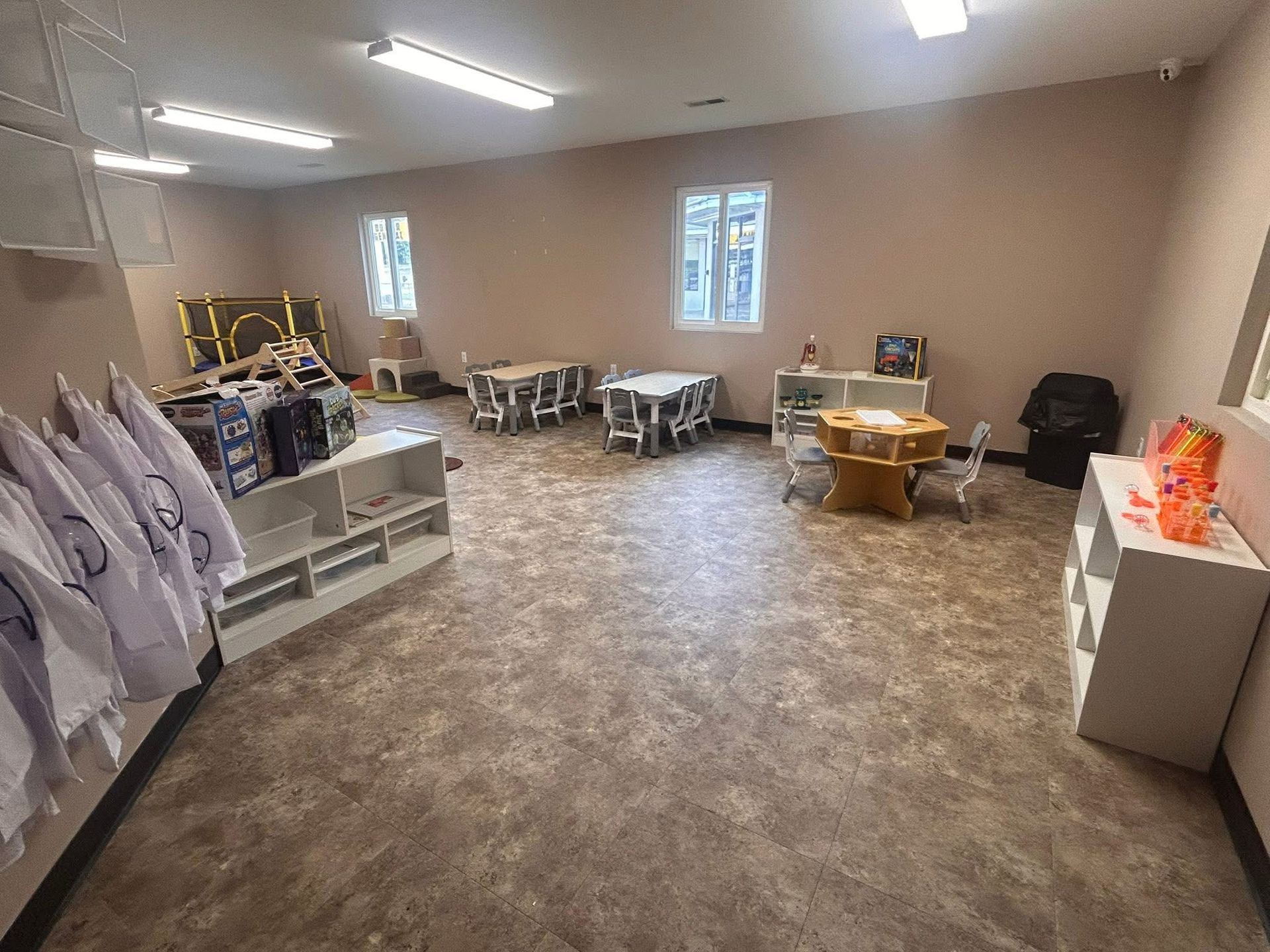 Empty classroom with tables, shelves, lab coats hanging on left wall, windows along back wall.