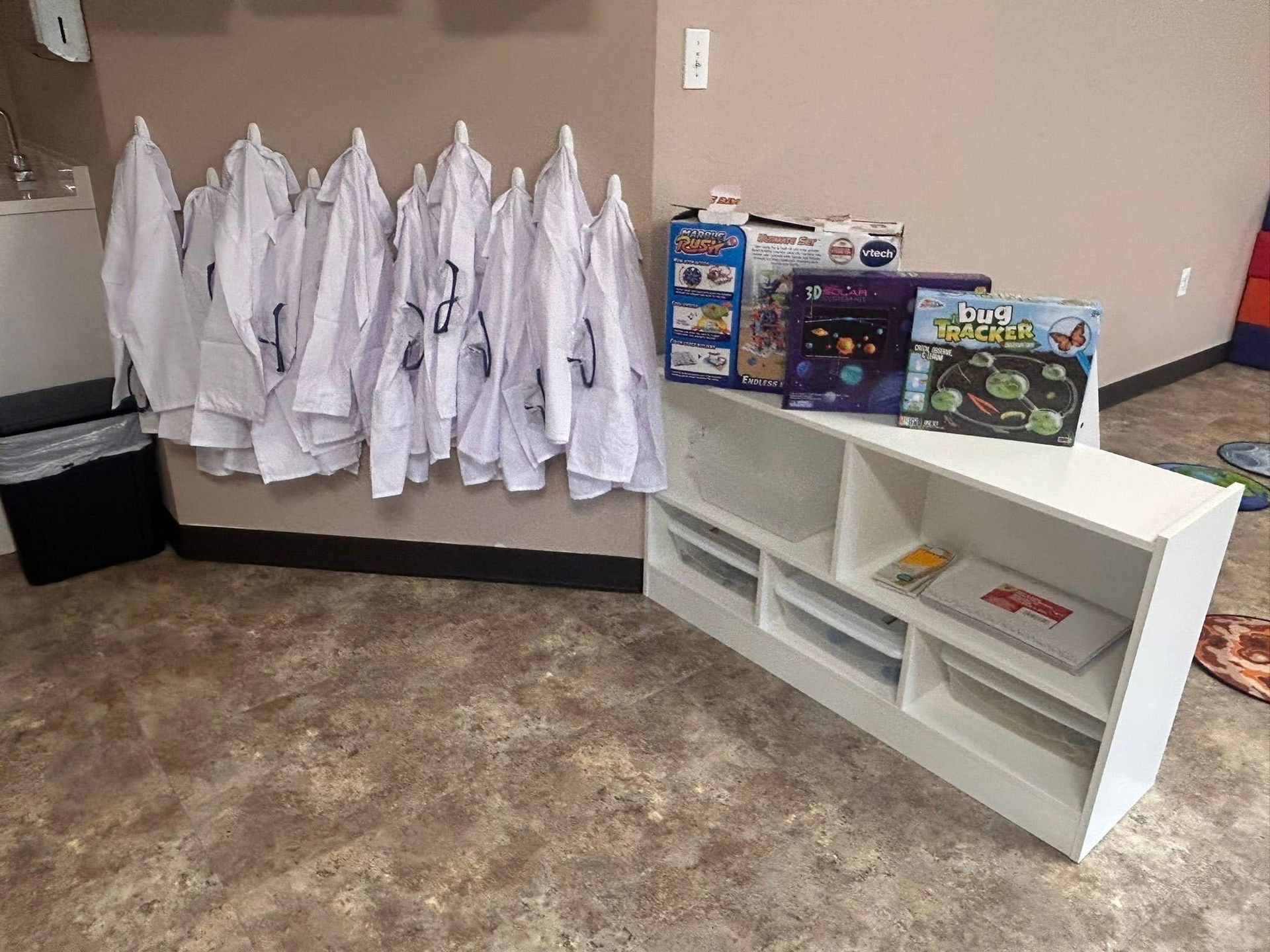 White lab coats hang on hooks. White shelf with boxes and games.