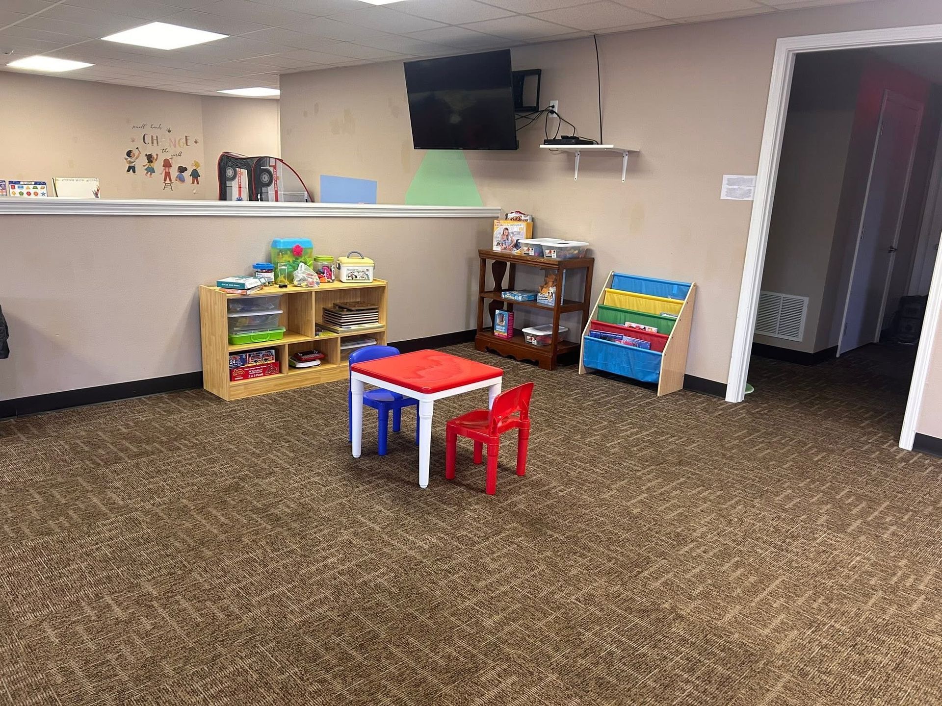 A playroom with a red table, blue and red chairs, toys, and a TV on a beige wall.