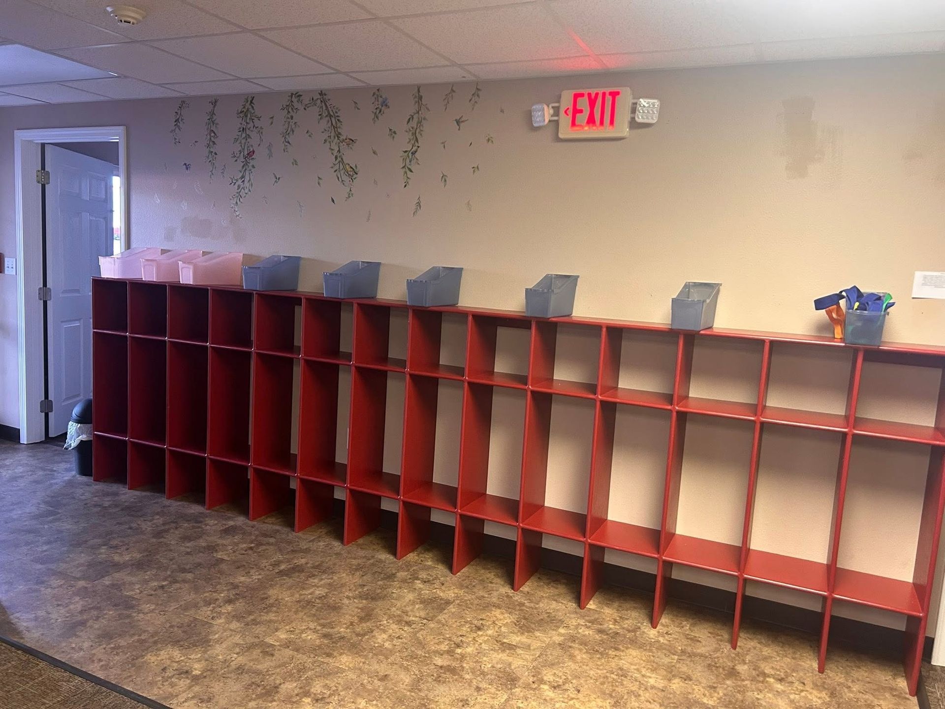 Red cubbies line a wall; gray bins sit atop. An exit sign is above.