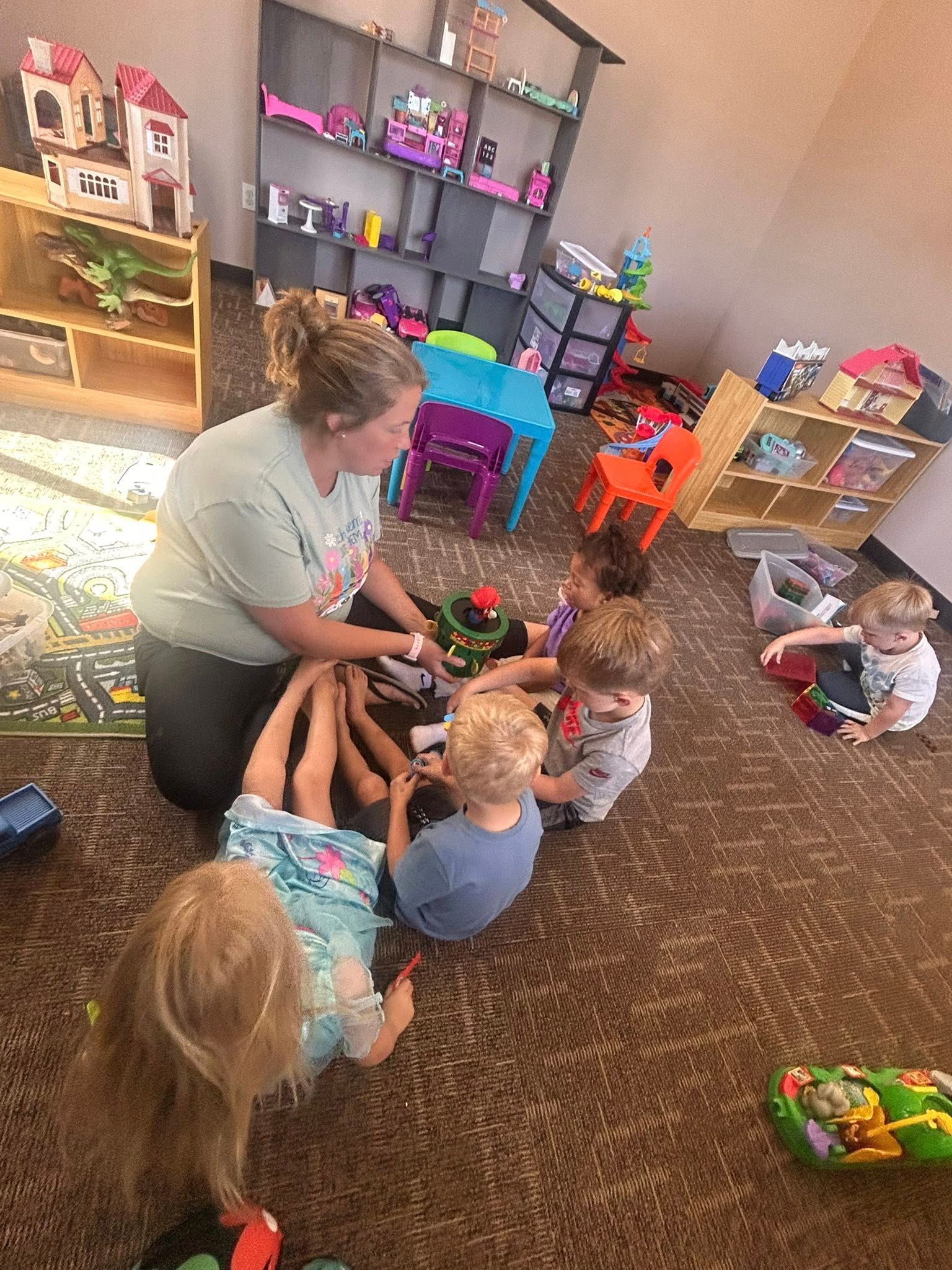 Woman playing with toys with five children on a carpeted floor in a colorful playroom.
