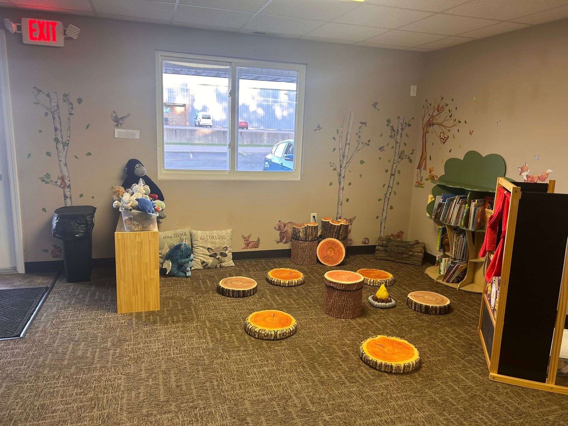 Cozy children's reading area. Features tree stump seats, a bookshelf, and forest-themed wall art.