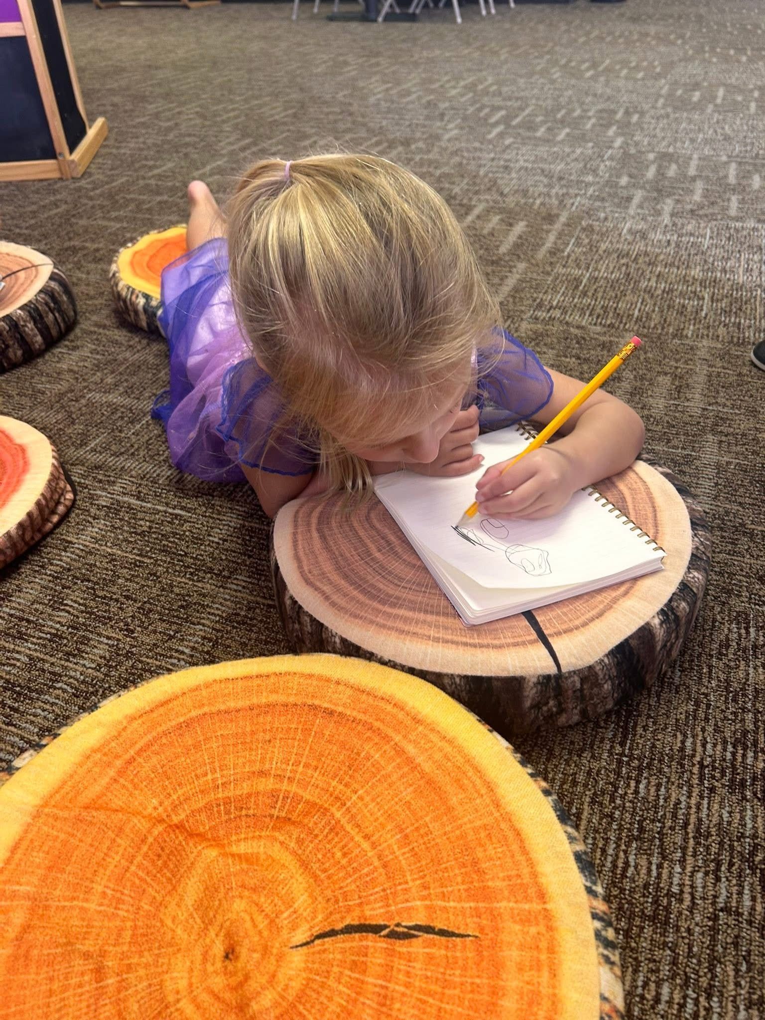 A young girl in a purple dress lies on a rug, drawing in a notepad, using a pencil, with tree stump cushions.