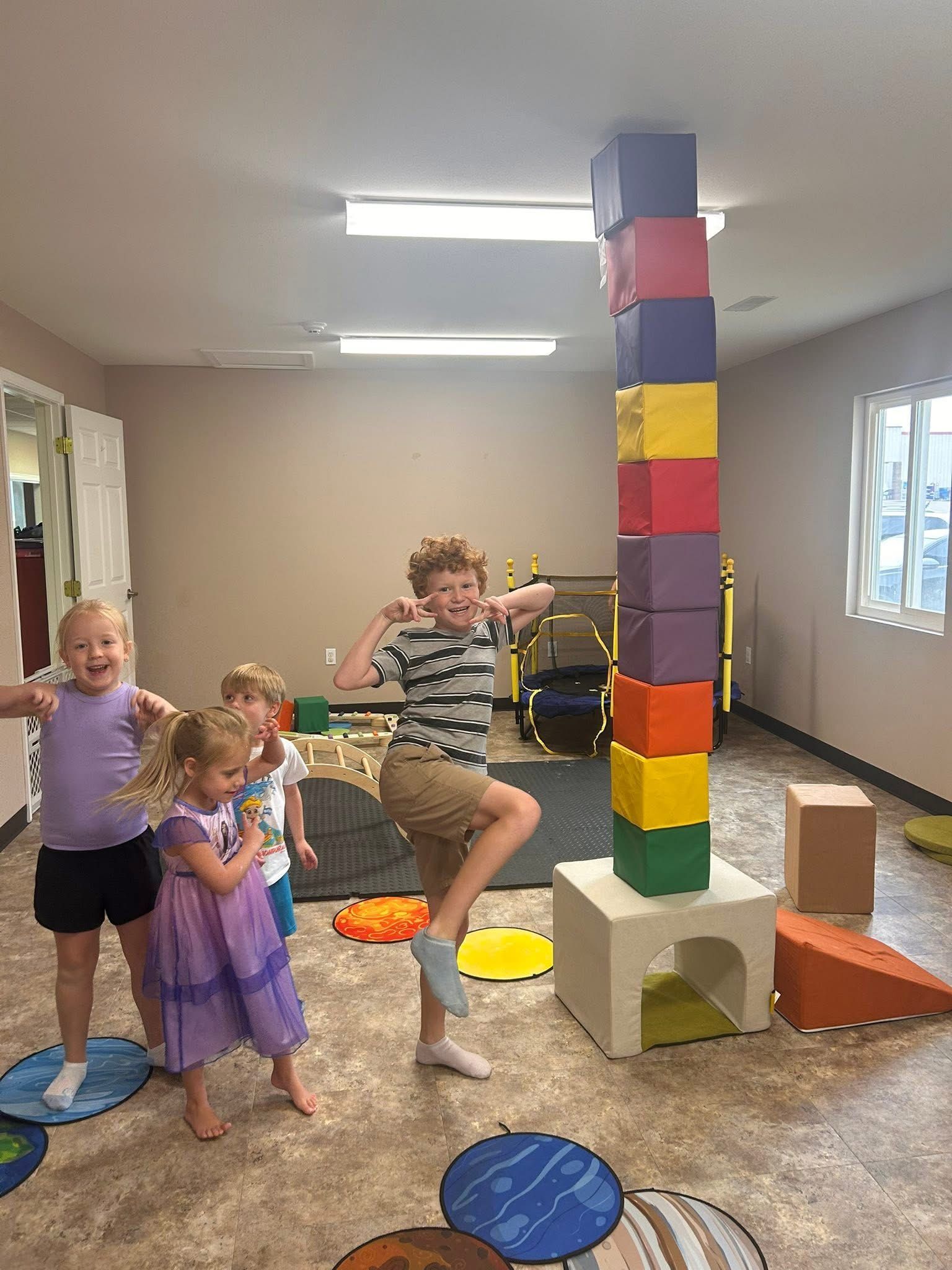 Children around a tall tower of colorful blocks in a play area. One boy poses, others smile.