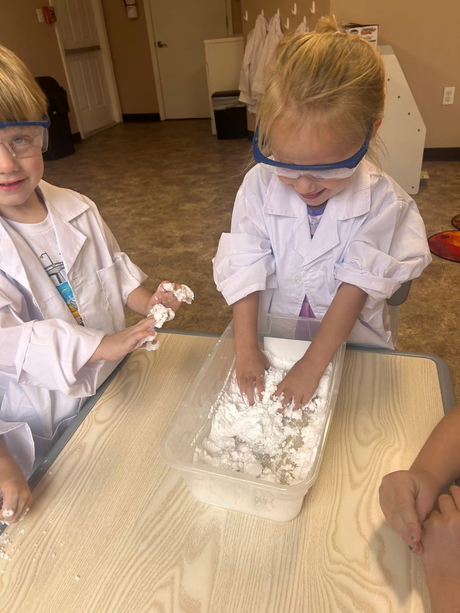 Two children in lab coats and goggles play with a white, powdery substance in a container.