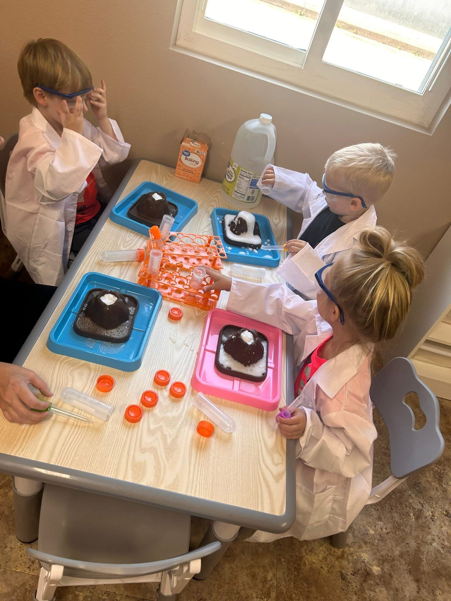Four kids in lab coats and safety glasses doing a science experiment at a table, with trays and baking soda.