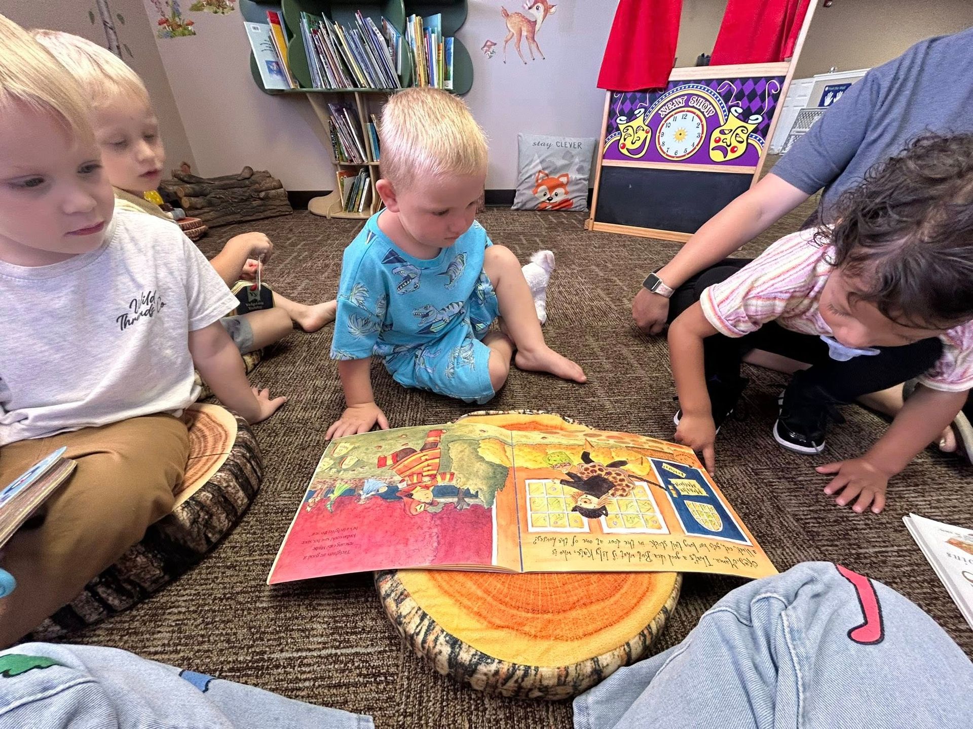 Children reading a book on a floor with tree stump cushions, in a library setting.