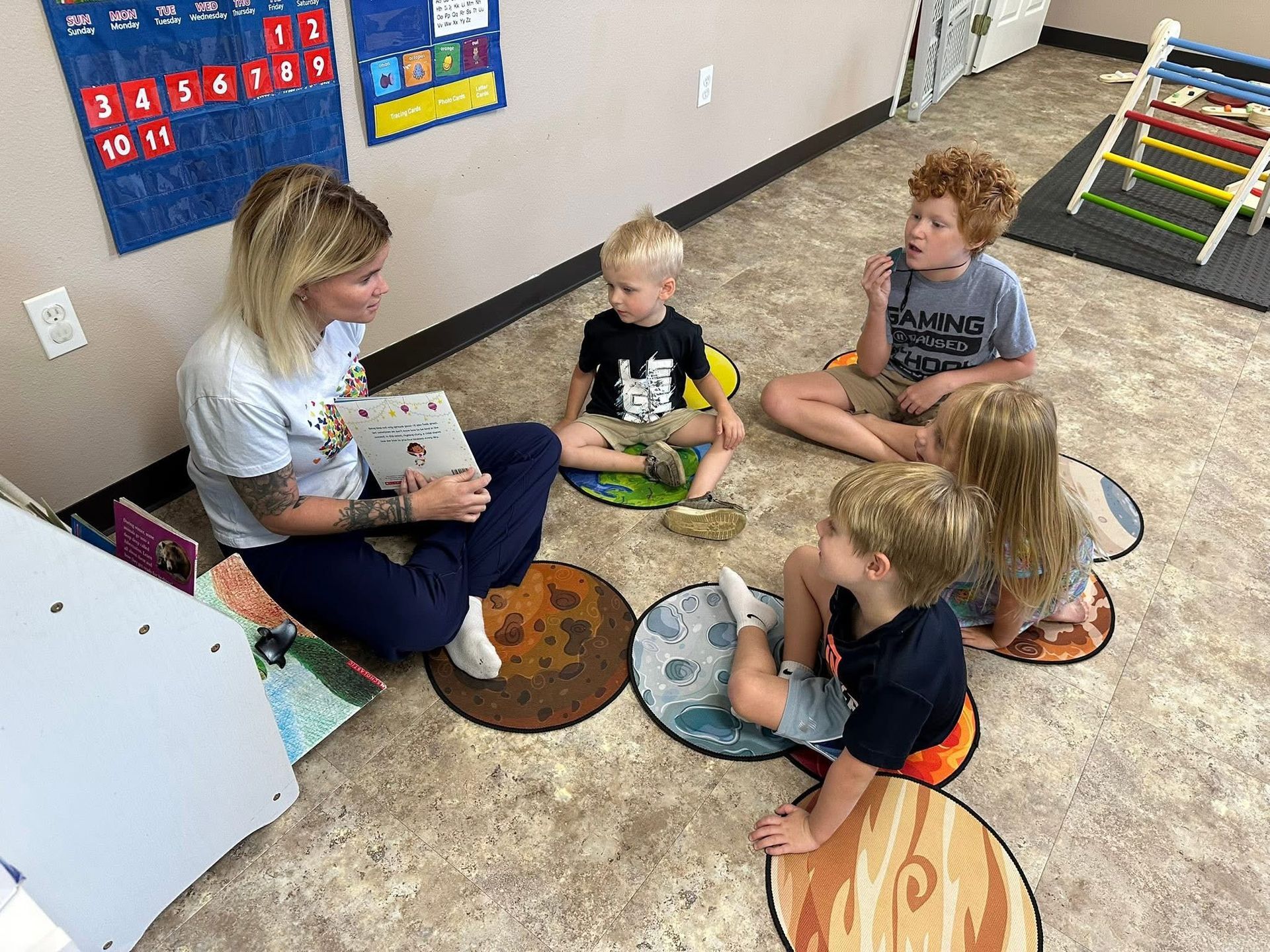 Teacher reads a book to a group of children sitting on planet-themed floor cushions.