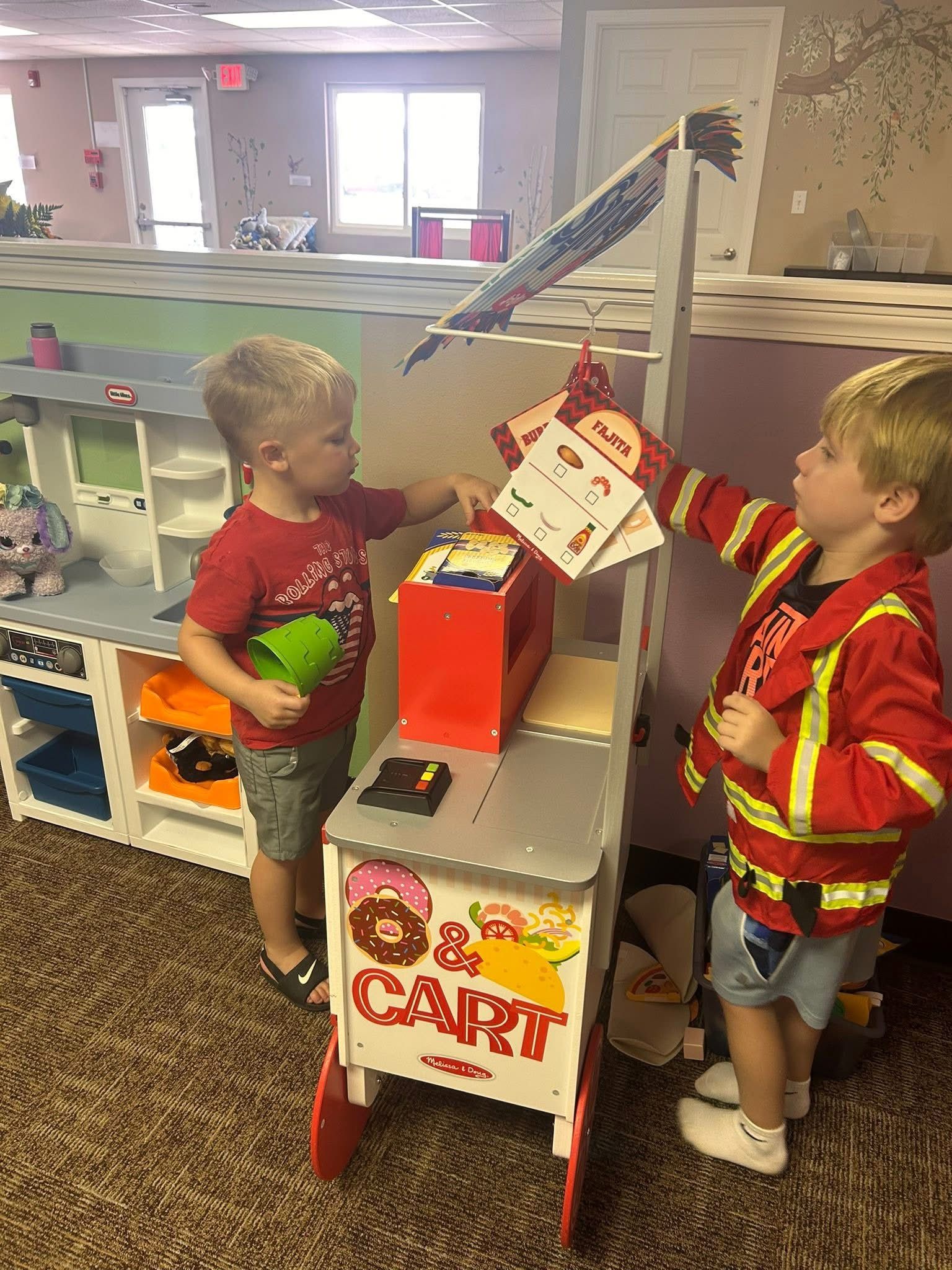 Two young boys playing with a toy food cart, one holding a menu, the other a cup.