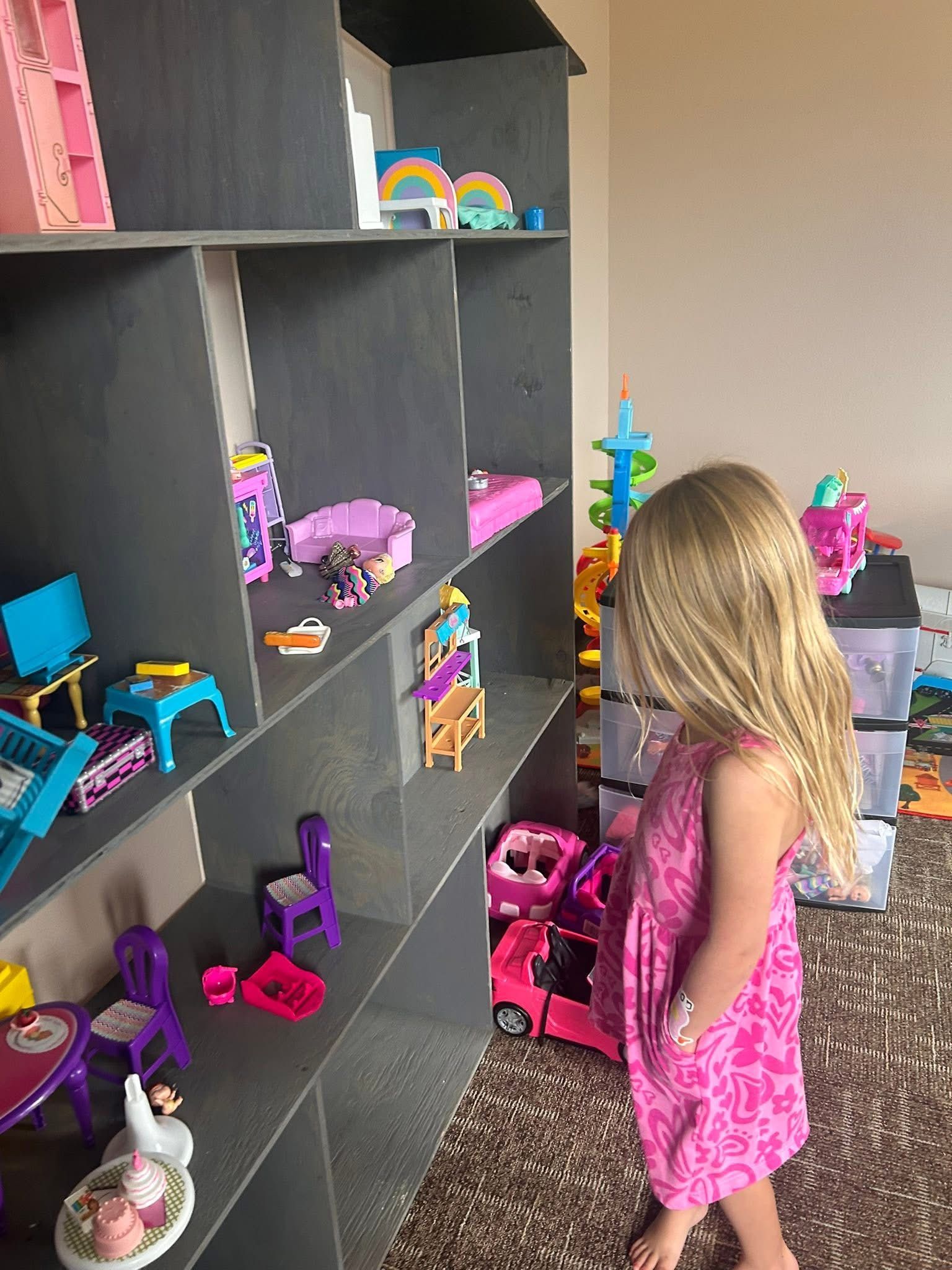 A young girl in a pink dress looks at a large dollhouse with colorful furniture and toys.
