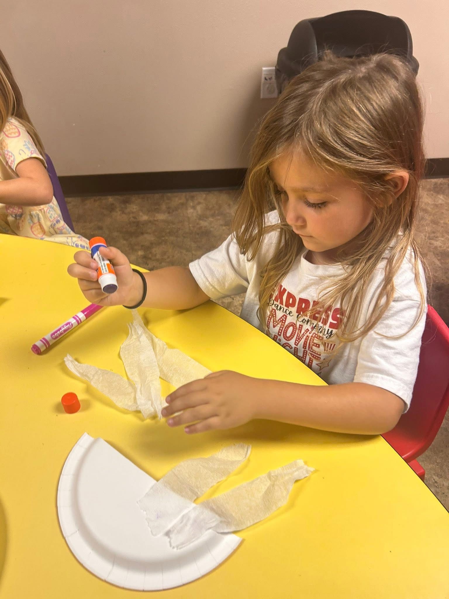Girl gluing fabric to a paper plate at a yellow table.