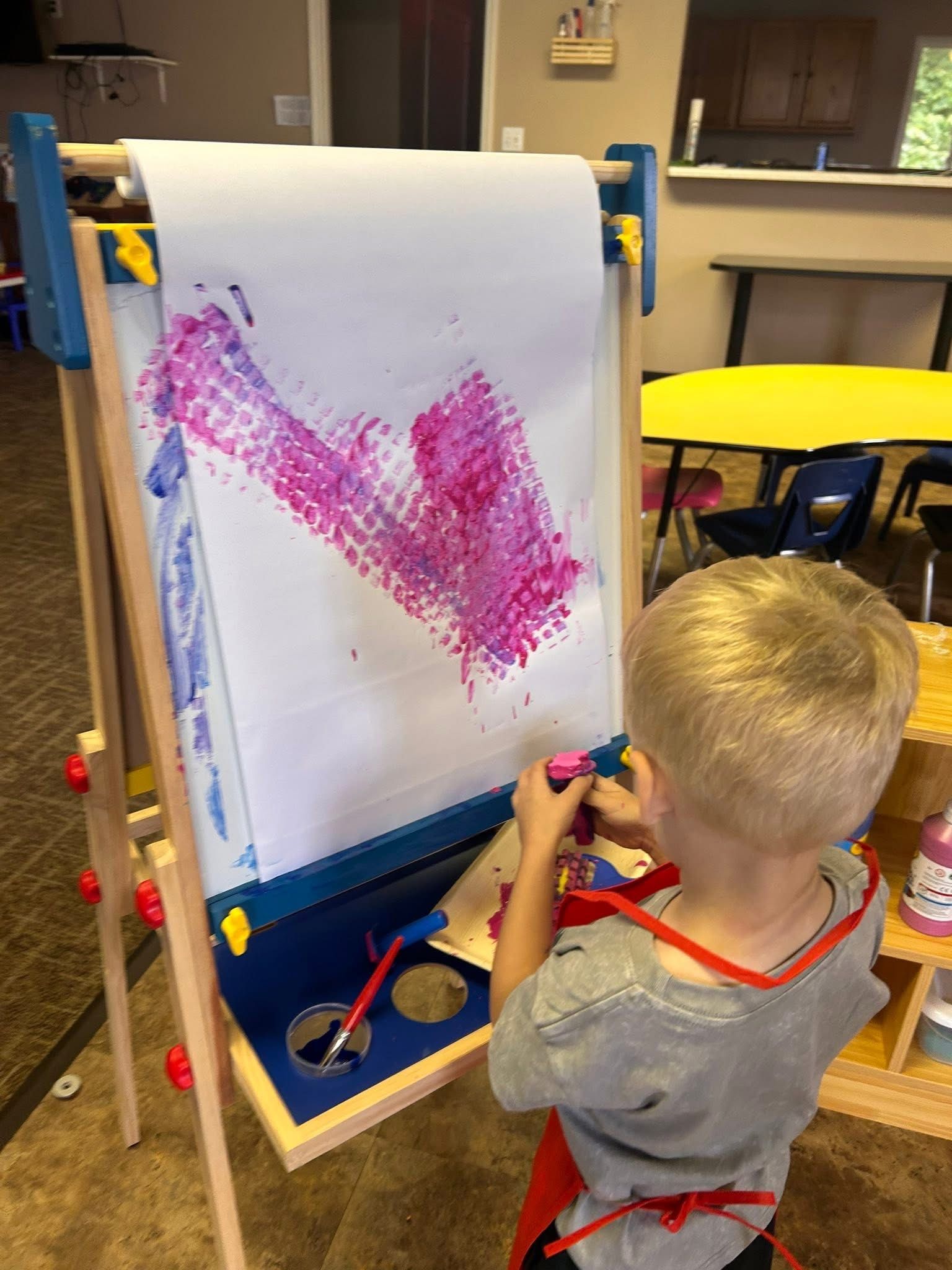 Blond child painting with pink paint on an easel in a classroom.