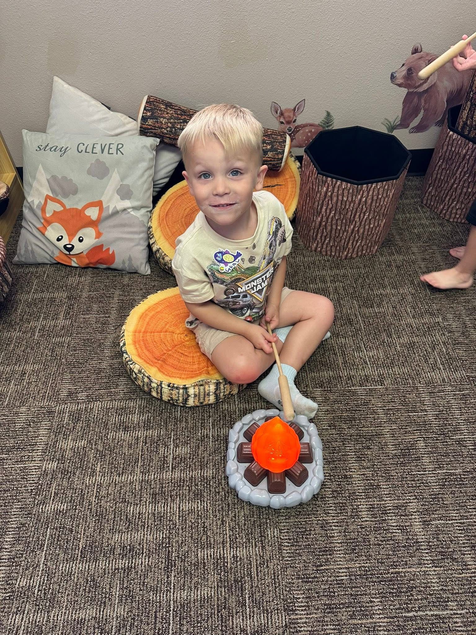 Blond boy sits on wooden slices near a toy campfire, smiling. Indoors with wood and fox décor.