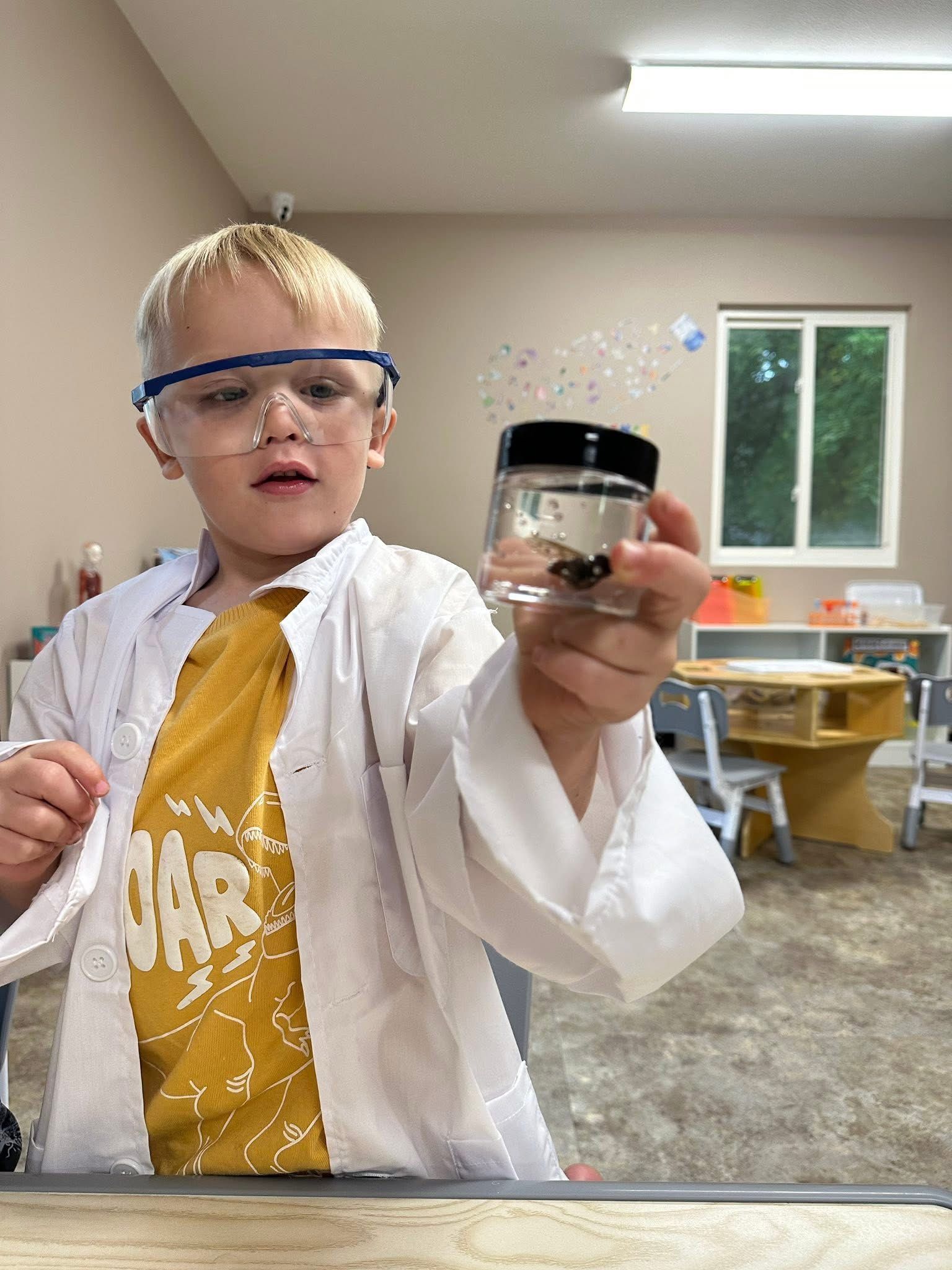 Young boy in a lab coat and goggles examining a jar, in a classroom.