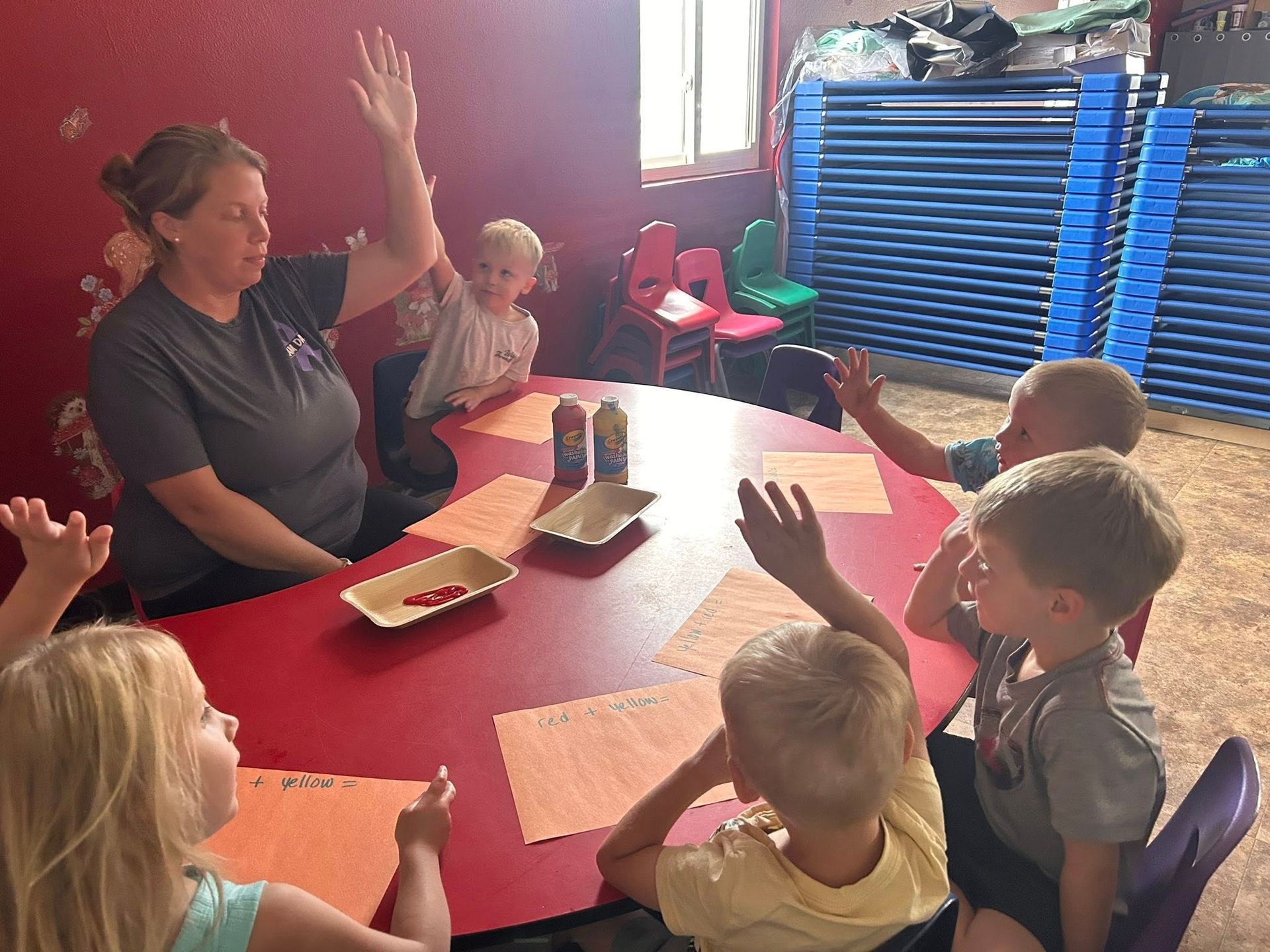 Teacher and children at a round table with hands raised in a classroom setting.